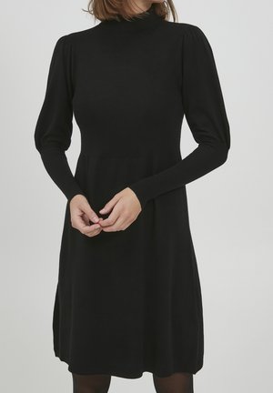 Kobieta ubrana w czarną sukienkę z długim rękawem i wysokim kołnierzem z bufkami na ramionach, stojąca z rękami luźno złączonymi na wysokości talii.