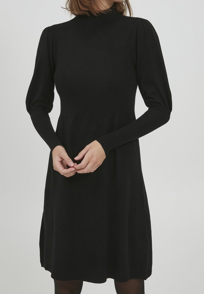 Femme portant une robe noire à manches longues et col montant, avec des épaules bouffantes, debout les mains légèrement jointes au niveau de la taille.