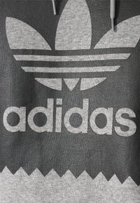 Felpa grigia con un grande logo Adidas Trefoil bianco e testo. Presenta un tessuto strutturato e un design a zigzag del fondo a contrasto.