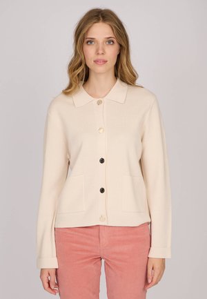 Femme portant un cardigan crème à boutons avec deux poches avant et un pantalon en velours côtelé rose, se tenant devant un fond clair.