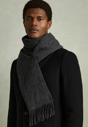 Mees, kes kannab musta mantlit ja tumedat halli tuttidega salli, seisab otse vaataja poole tavalise heleda tausta ees.