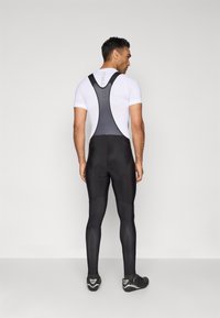 Sorte sykkelbuks med nettingstropper, med en tettsittende design og teksturert stoff. Vist bakfra, sammen med sorte sykkelsko.