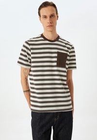 Stribet t-shirt i beige og brun bomuld. Ribbet rund hals, korte ærmer, med en brun brystlomme, struktureret stof.