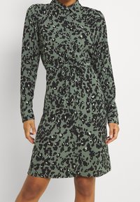 Klänning i leopardmönster i olivgrön, med krage, långa ärmar, knytning i midjan och knappstängning. Slät tygtextur.