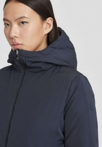 Marineblauwe pufferjack met een hoge kraag en een rits. Beschikt over een klein logo op de mouw. Soepele, matte textuur.