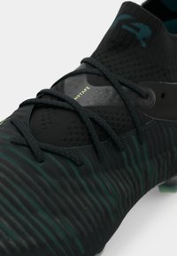 Botín de fútbol negro con diseño de rayas verdes, que presenta un upper texturizado y cordones negros. Incluye un sutil logo en el collar del tobillo.