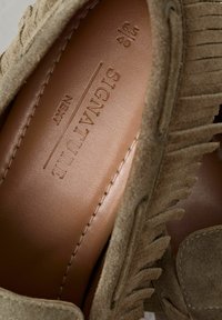 Mocasines de ante en beige terroso, con un detalle de flecos, forro de cuero suave y marca estampada en la plantilla.