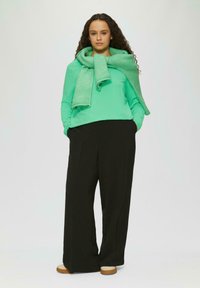 Groene blouse met lange mouwen, gecombineerd met zwarte wijde broek. Een bijpassende groene gebreide sjaal hangt casual om de nek. Minimalistisch ontwerp.