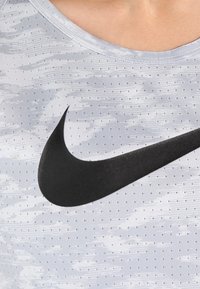 Lichtgrijze sporttop met een gestippelde textuur, met een opvallend zwarte Nike Swoosh-logo. Ronde halslijn, korte mouwen.