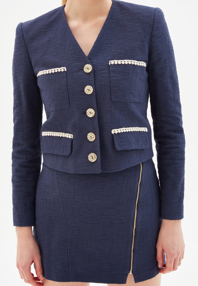 Blazer texturé bleu marine avec col en V, deux poches avant et cinq boutons décoratifs. La jupe possède une fermeture éclair latérale et est en tissu assorti.