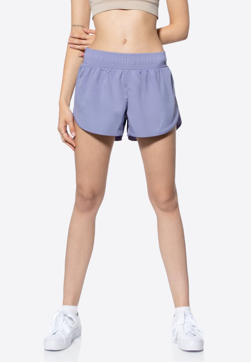 Shorts de sport violet clair avec une taille élastique, présentant un ourlet courbe et des fentes latérales pour une meilleure mobilité. Associés à des baskets blanches.