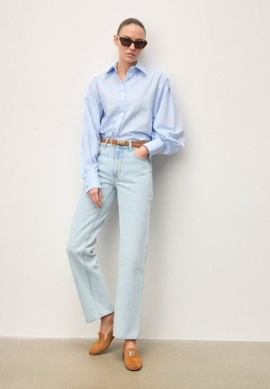 Hellblaues oversize Hemd, hoch taillierte helle Denim-Jeans, brauner Gürtel und beigefarbene Wildleder-Mules mit goldfarbenem Akzent. Glatte Texturen, lockere Passform.