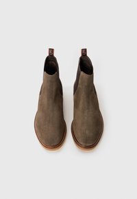 Un par de botas Chelsea de ante marrón con paneles laterales elásticos y tiradores sobre un fondo blanco.