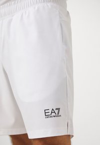 Calções atléticos brancos com uma faixa elástica na cintura, bolsos laterais e um logo preto EA7 Emporio Armani na parte inferior da perna esquerda. Textura de tecido suave.