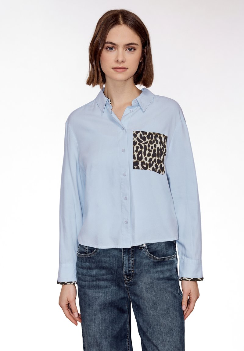 Femme portant une chemise à manches longues bleu clair avec un détail de poche et de poignets à imprimé léopard, associée à un jean bleu, debout devant un fond blanc.