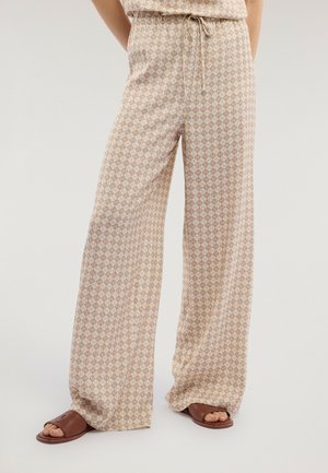 Beige og hvide bukser med vidde i benene og diamantmønster, med snørelukning i taljen, båret med brune åbne slide-sandaler på neutral baggrund.