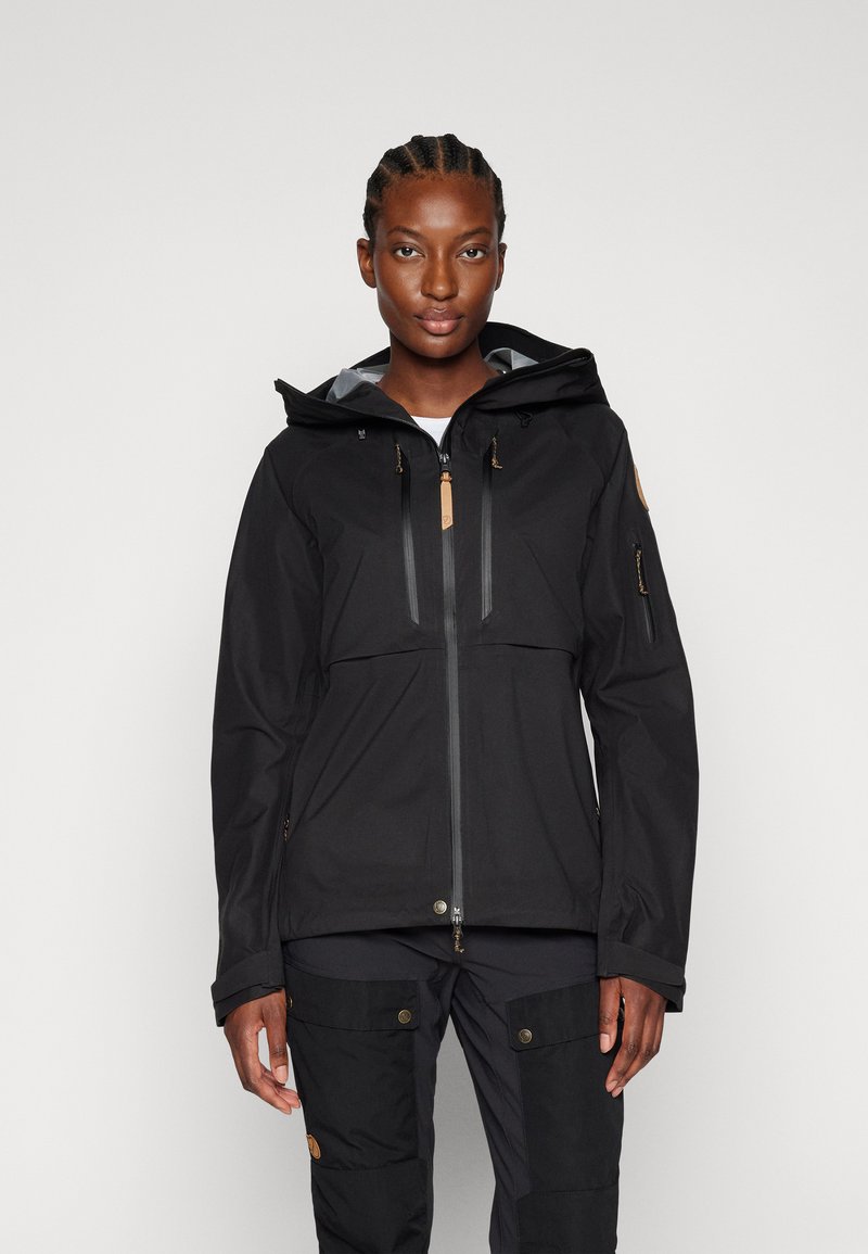 Veste noire imperméable avec une fermeture éclair sur le devant, une capuche ajustable et deux poches poitrine. Affiche une texture lisse et un branding discret.