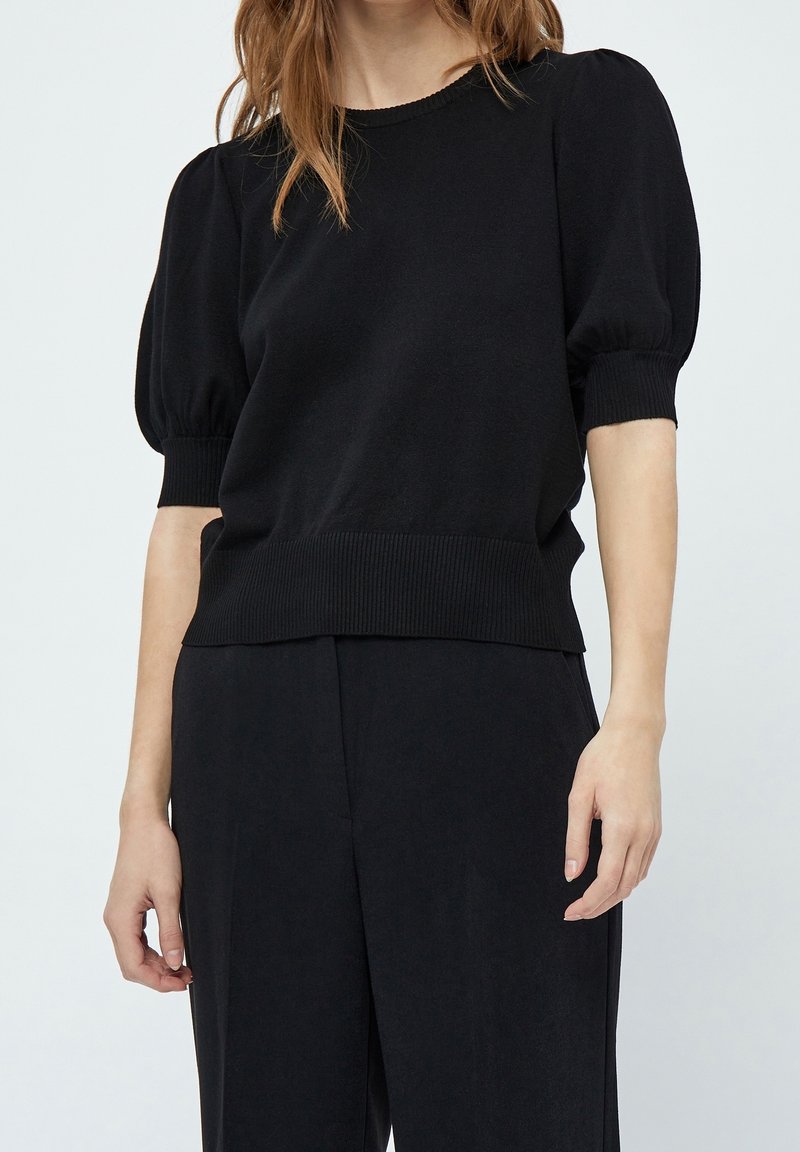 Žena v černém topu s krátkým rukávem a našitými rukávy, doplněná o černé kalhoty s širokými nohavicemi, na jednobarevném pozadí.