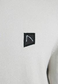 Lichtgrijze gebreide trui met een klein zwart patch en een witte grafische print. De stof is zacht met een fijne textuur.