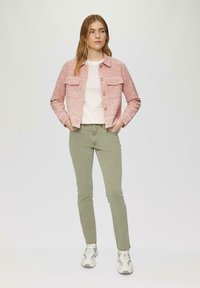 Rood-wit geruite jas met twee voorzakken, witte t-shirt met tekst en olijfgroene slim-fit broek. Zilveren sneakers maken de outfit compleet.
