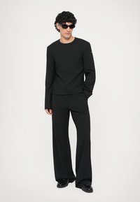 Mann in einem kompletten schwarzen Outfit mit langärmeligem Oberteil, weit geschnittenen Hosen, schwarzen Schuhen und dunkler Sonnenbrille, der mit einer Hand in der Tasche steht.