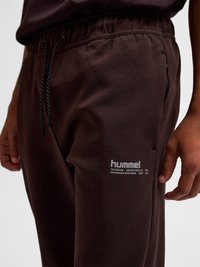 Nahaufnahme einer Person, die dunkelbraune Jogginghosen mit Kordelzug trägt, mit „hummel“-Logo und Reißverschlusstasche an der rechten Oberschenkelseite.