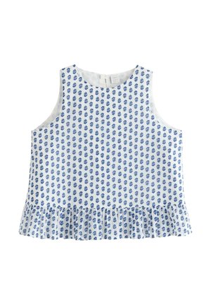 Ein lächelndes Mädchen steht und trägt ein ärmelloses weißes Oberteil mit blauem Muster, Jeansshorts, weiße Socken und weiße Sneakers vor einem einfarbigen Hintergrund.