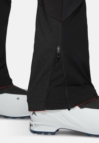 Zwarte sportbroek met een textuurpaneel, ritssluitingen aan de zijkanten en rode accenten, gecombineerd met witte sportschoenen met blauwe zolen.