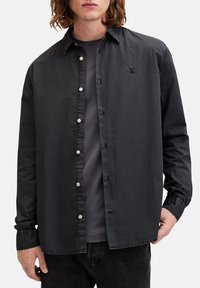 Camisa negra de manga larga con botones, de textura suave, que presenta un sutil bordado de logo y botones de color claro en el frente.