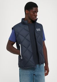 Homme portant un gilet matelassé marine sur un t-shirt bleu foncé et un jean bleu clair, avec une main dans la poche du gilet, regardant vers le bas.