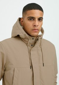 Beige windbreaker jacket with a hood, featuring a front zip closure, adjustable drawstrings, and a smooth, textured fabric.