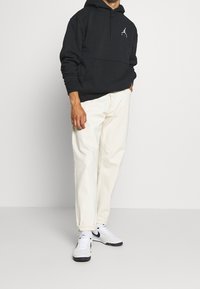 Felpa nera con tasca a marsupio e logo bianco, abbinata a pantaloni bianchi larghi e sneakers bianche con dettagli neri.