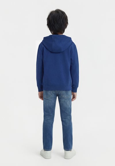 Αγόρι που στέκεται γυρισμένο πλάτη, φοράει μπλε φούτερ με κουκούλα, μπλε τζιν και λευκά αθλητικά παπούτσια, μπροστά σε απλό λευκό φόντο.
