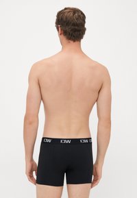 Giovane uomo visto di spalle, che indossa boxer neri con una fascia in vita bianca con il logo "ICANIWILL" su sfondo bianco.