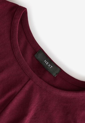 Top in cotone color bordeaux con scollo arrotondato, dotato di un'etichetta nera con la scritta "NEXT." Texture liscia e dettagli cuciti visibili.