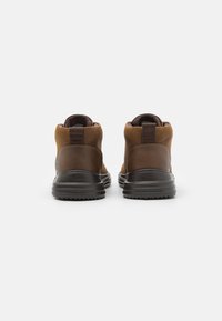 Botas de tobillo de cuero marrón con suela de goma negra, cuello acolchado y tirador en la parte trasera. Se aprecian superficies lisas y texturizadas.