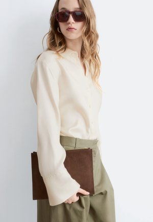 Vrouw met golvend blond haar die oversized bruine zonnebrillen draagt, een crèmekleurige blouse, olijfgroene broek en een bruine clutch vasthoudt.