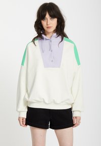 Weißer, übergroßer Sweatshirt mit einem lila Panel und grünen Akzenten, ausgestattet mit einem hohen Kragen, Knöpfen und strukturierten Ärmeln. Schwarze Shorts runden den Look ab.