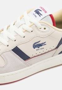 Lacoste sneakers met een beige leren bovenkant, marineblauwe accenten en een suède overlay. Beschikt over een rode hiel detail en een logo dat aan de zijkant is gedrukt.