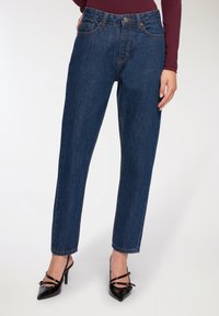 Wąskie niebieskie dżinsy носzone z czarnymi szpilkami w kształcie spiczastym oraz długim rękawem burgundową bluzką, pokazane od talii do stóp.