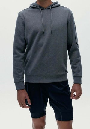 Person iført en mørkegrå hættetrøje med snore og sorte sportsshorts, stående mod en ensartet lys baggrund.