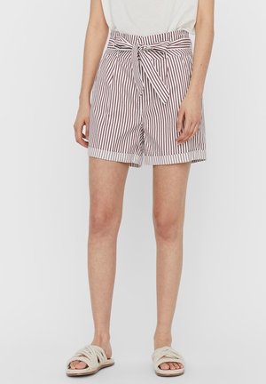Kvinde iført hvide shorts med høj talje og lodrette brune striber samt et bundet bælte, kombineret med hvide reb-sandaler og en hvid top.