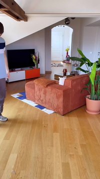 Vyras stovi ryškiai apšviestame svetainėje su oranžine sofa, dideliu žaliuoju augalu, televizoriumi ant žemo kabineto ir šiuolaikiniais mediniais grindimis.