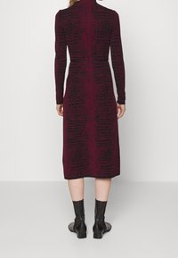Femme portant une robe noire à manches longues et longueur moitié-mollet, avec un motif abstrait rouge foncé, associée à des bottines noires, se tenant de dos.