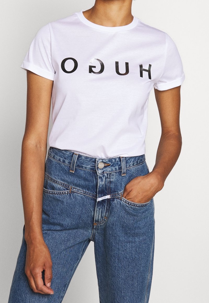 Camiseta blanca de manga corta con un logo impreso en negro "HUGO" en el pecho, combinada con unos vaqueros de denim azul de tiro alto.