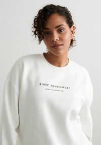 Biely sveter s okrúhlym výstrihom, rebrovanými manžetami a potlačou "Aim'n Sportswear" v čiernej farbe. Hladká textúra, voľný strih, ľahký materiál.