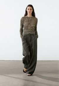 Strickoberteil mit strukturiertem Muster in neutralen Tönen, kombiniert mit lockeren grauen Hosen und schwarz-gestrickten Sandalen. Auffällige lange Halskette als Akzent.