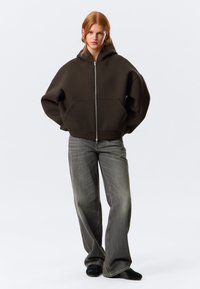 Sweatshirt verde oliva escuro com capuz, fecho de correr e corte oversized, com bolsos à frente e punhos canelados, combinado com jeans claros de perna larga.