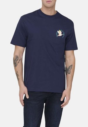 Homme portant un T-shirt à manches courtes bleu marine avec un petit logo d'oiseau coloré sur le côté gauche de la poitrine et un jean foncé, tatouages visibles sur les deux bras.