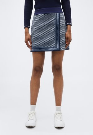 Vrouw draagt een marineblauwe wikkelrok met patroon, witte sokken, witte sneakers en een marineblauwe top met lange mouwen en witte afwerking aan de manchetten.